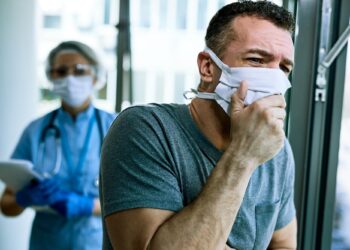 Maske takan öksüren bir adam ve arkasında tıbbi kıyafetli sağlık çalışanı.