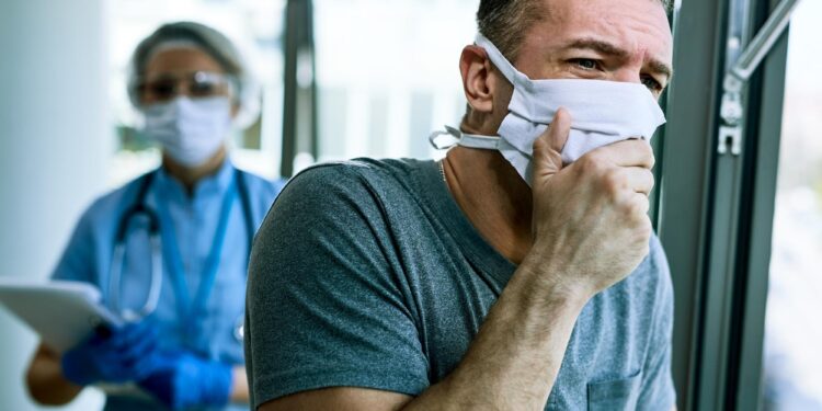 Maske takan öksüren bir adam ve arkasında tıbbi kıyafetli sağlık çalışanı.