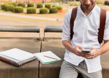 Beyaz gömlekli bir öğrenci açık havada oturup akıllı telefon kullanıyor.