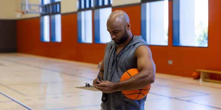 Antrenörlük kıyafeti giymiş, elinde basketbol topu ve pano tutan koç.