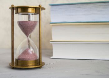 Masada duran pembe kumlu antika bir kum saati ve arkasında üst üste dizilmiş kitaplar.