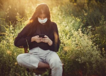 Doğada çimenler arasında oturup maske takarak cep telefonu kullanan kadın.