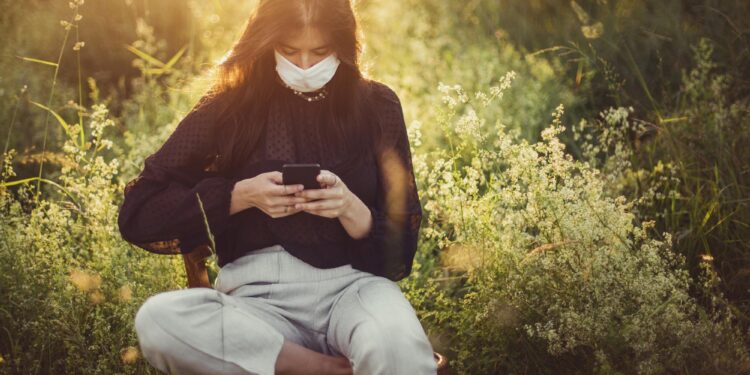 Doğada çimenler arasında oturup maske takarak cep telefonu kullanan kadın.