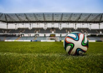 Çim sahada duran renkli bir futbol topu ve arkasında büyük stadyum tribünleri var.
