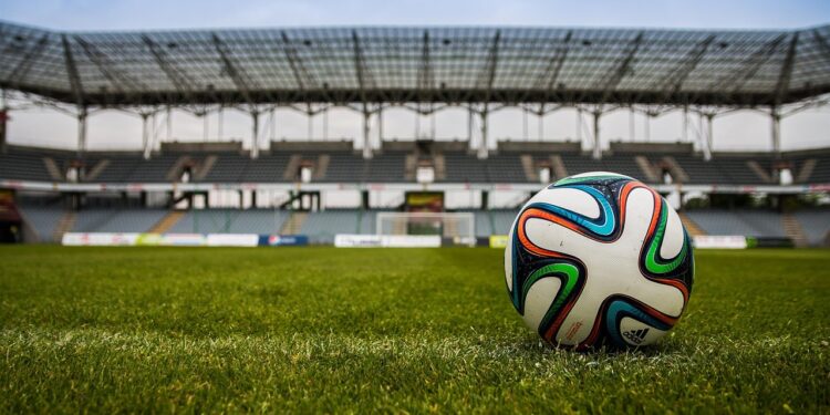 Çim sahada duran renkli bir futbol topu ve arkasında büyük stadyum tribünleri var.