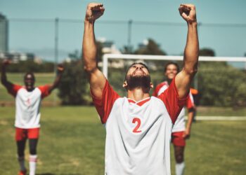 Kollarını havaya kaldırarak sevinç çığlığı atan sporcu ve arka planda takım arkadaşları.
