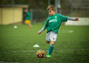 Futbol sahasında yeşil formayla topa vuran küçük çocuk oyuncu.