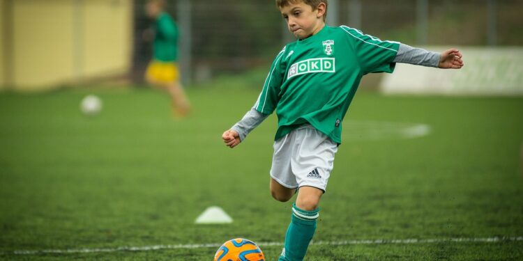 Futbol sahasında yeşil formayla topa vuran küçük çocuk oyuncu.