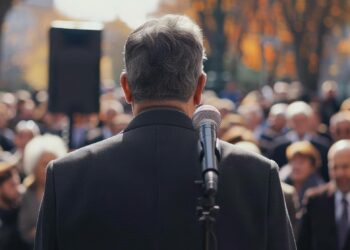 Mikrofon önünde duran, kalabalığa hitap eden bir siyasetçinin sırtı.