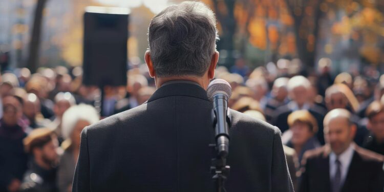 Mikrofon önünde duran, kalabalığa hitap eden bir siyasetçinin sırtı.
