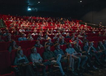 Kırmızı koltuklu sinema salonunda film izleyen kalabalık izleyiciler ve dev perde