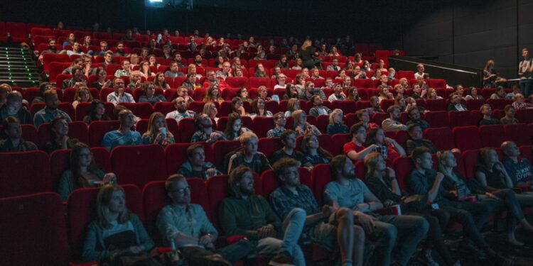 Kırmızı koltuklu sinema salonunda film izleyen kalabalık izleyiciler ve dev perde