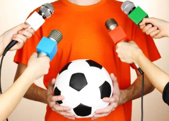 Turuncu tişört giyen bir kişinin etrafını saran mikrofonlar ve futbol topu.