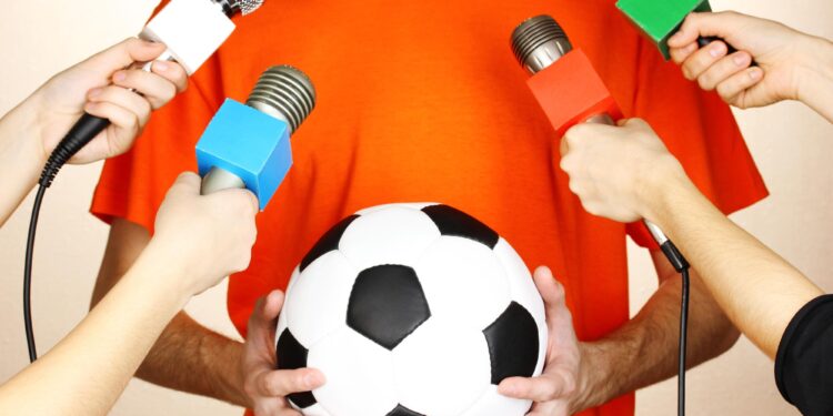 Turuncu tişört giyen bir kişinin etrafını saran mikrofonlar ve futbol topu.