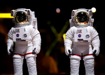 Karanlık arka planda duran iki adet beyaz astronot kıyafeti ve kaskı.