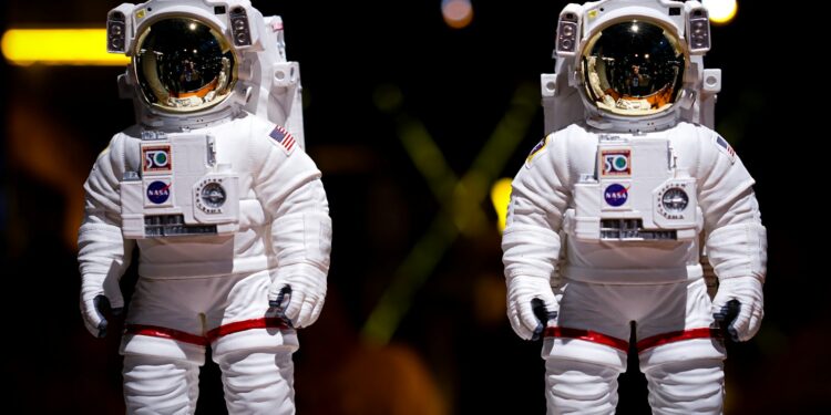 Karanlık arka planda duran iki adet beyaz astronot kıyafeti ve kaskı.